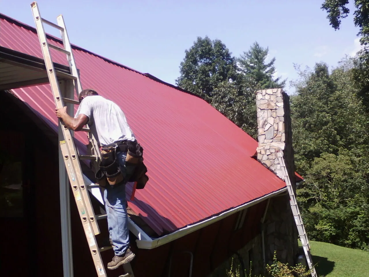 Roofer on a ladder beside a red metal roof and stone chimney — Soffit & Fascia Repair by Roofing Brothers in Clemons